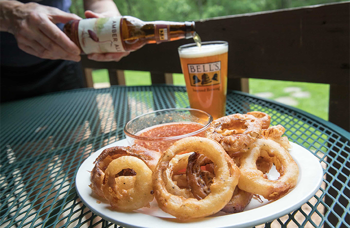 A plate of onion rings with orange dipping sauce in a bowl, with a bottle of Amber Ale being poured into a pint glass in the background