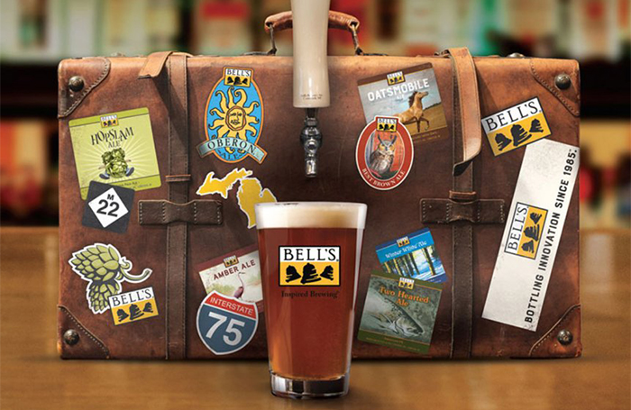 A glass of beer in front of a suitcase covered in travel stickers and bells logos with a tap attached to the front.