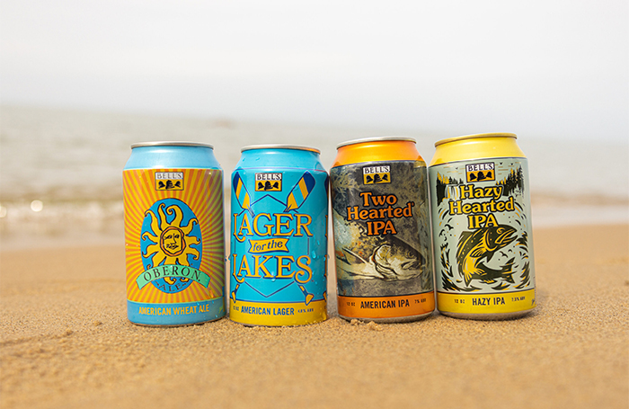 Four cans (Oberon, Lager for the Lakes, Two Hearted Ale and Hazy Hearted) on a beach with Lake Michigan in the background