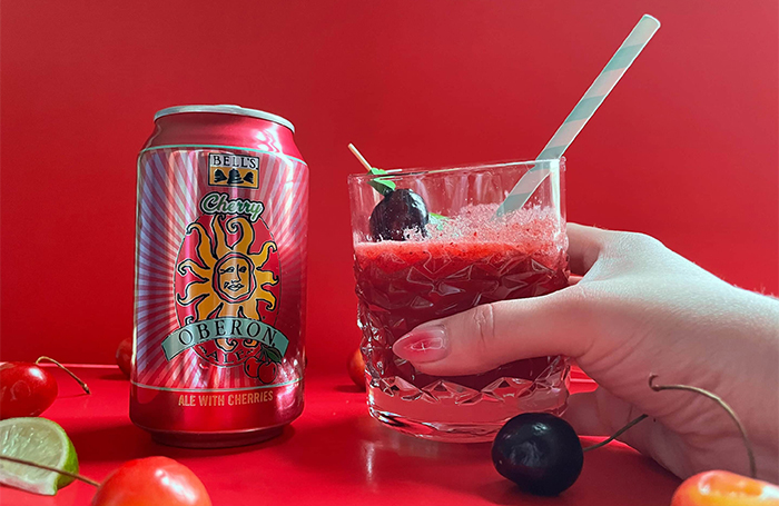 A bright red background with a can of Cherry Oberon and a glass of the red cherry Oberon Beer-rita, with cherries on the bottom of the picture. A hand is holding the beerita.