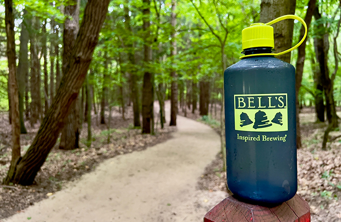 A water bottle with a Bell's logo next to a trail in the woods.