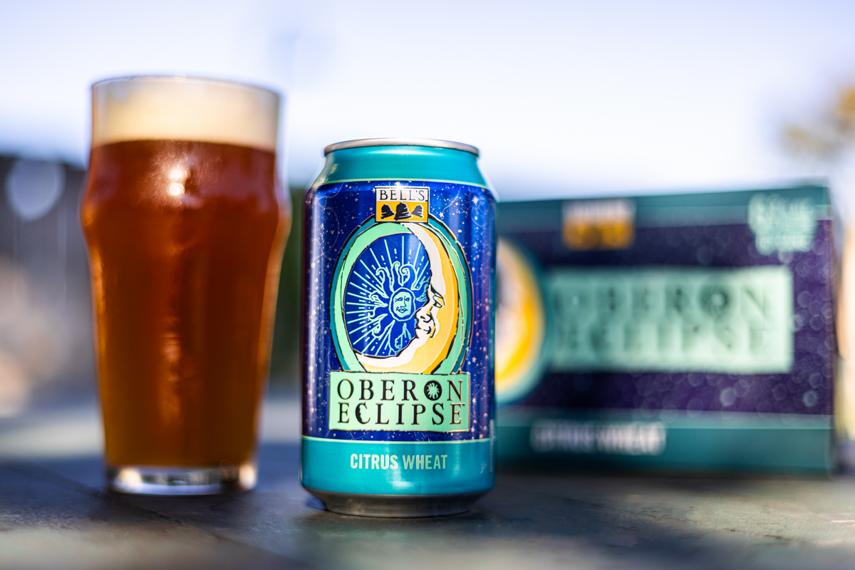 A can of Oberon Eclipse in front of a dark red-ish pint of beer and a six pack of beer