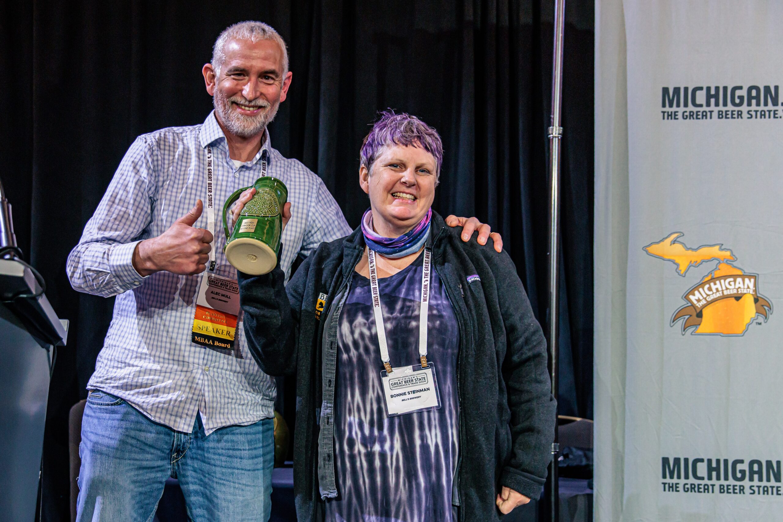 Bonnie and Alec stand smiling in front of a banner that reads "MICHIGAN THE GREAT BEER STATE." Alec is on the left, with gray hair and a beard, is holding a green beer stein trophy and giving a thumbs-up. The person on the right,Bonnie, with short purple hair, is wearing a black jacket.