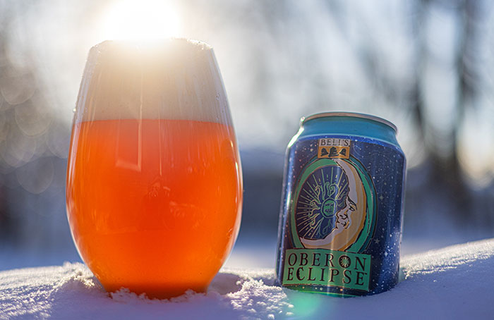 The image displays a vibrant, amber-colored beverage in a glass, set against a backdrop of a snowy landscape with sunlight filtering through. The glass is positioned on the snow, capturing the essence of a cold, bright winter's day. Next to the glass is a can of "Oberon Eclipse" beer from Bell's Brewery, which features a colorful design with a sun and moon motif, suggesting a limited edition or special brew. The overall scene evokes a sense of winter warmth, with the contrast between the cold environment and the inviting drink.