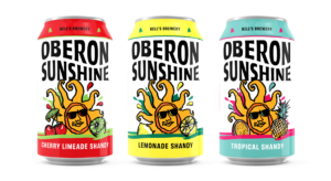 "Three cans of 'Oberon Sunshine' shandy from Bell's Brewery, each with a unique flavor. From left to right: 'Cherry Limeade Shandy' with a red tab and cherries and a lime depicted, 'Lemonade Shandy' with a yellow tab and a lemon slice, and 'Tropical Shandy' with a teal tab and a pineapple and orange slice. Each can has a sun with sunglasses in the center, varying background colors, and the Bell's Brewery logo at the top."