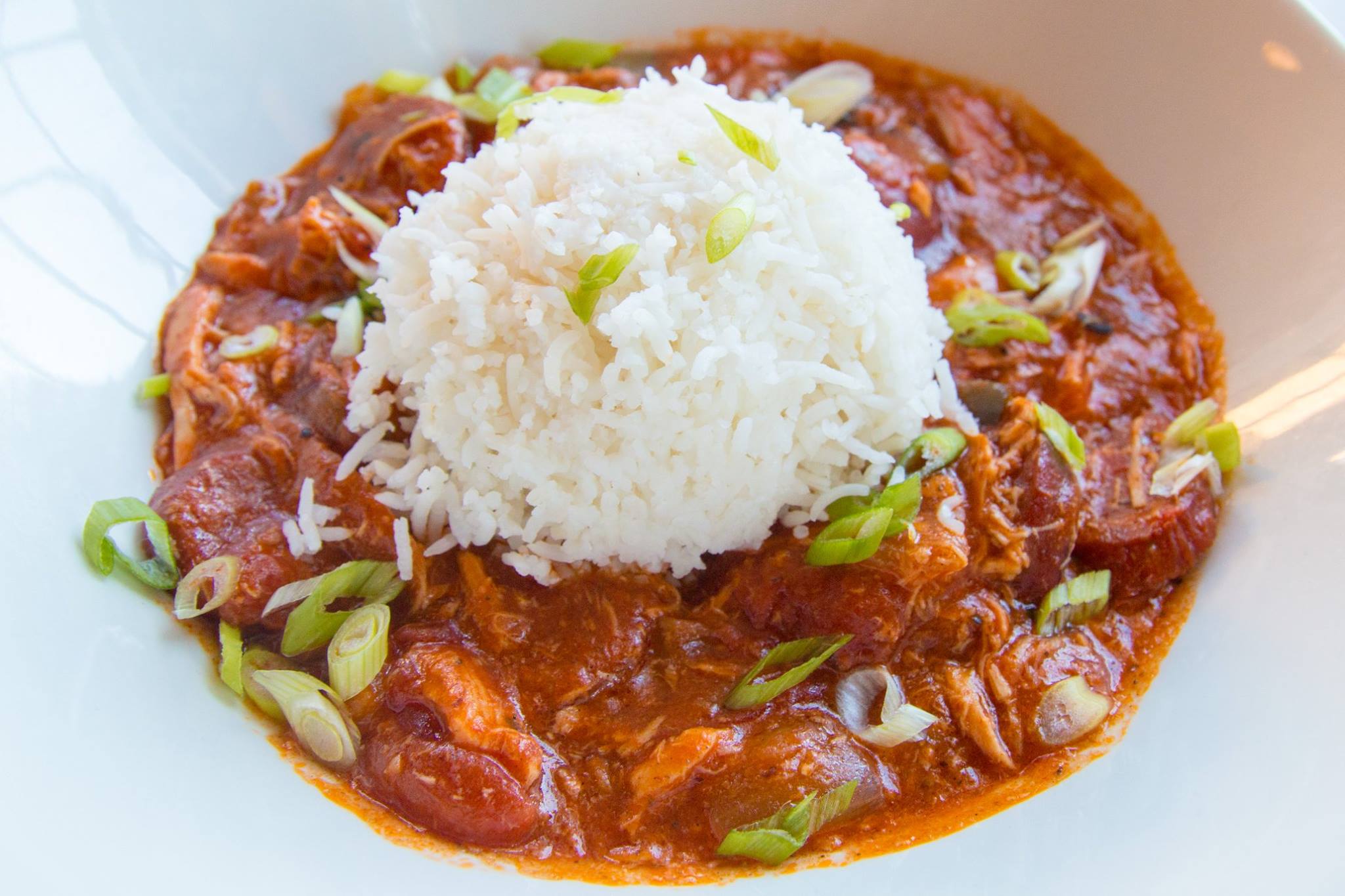 A plate of jambalaya topped with a mound of white rice, garnished with sliced green onions. The jambalaya has a rich red sauce with visible slices of Andouille sausage and hints of shredded chicken, indicative of a traditional, spicy Cajun dish.