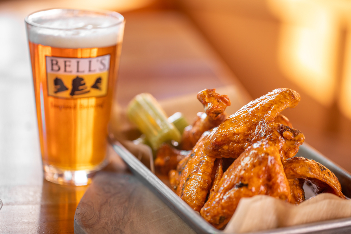 A pint of amber-colored beer in a branded Bell's glass, with crispy chicken wings coated in a glossy sauce, served with celery sticks on a metal tray. They are all on a wooden table.