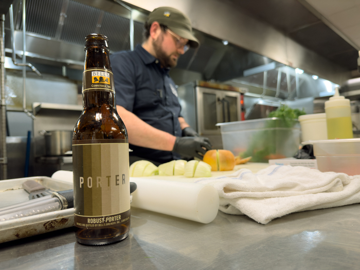 A bottle of Bell's Porter beer on a stainless steel kitchen counter with a chef cutting onions in the background.
