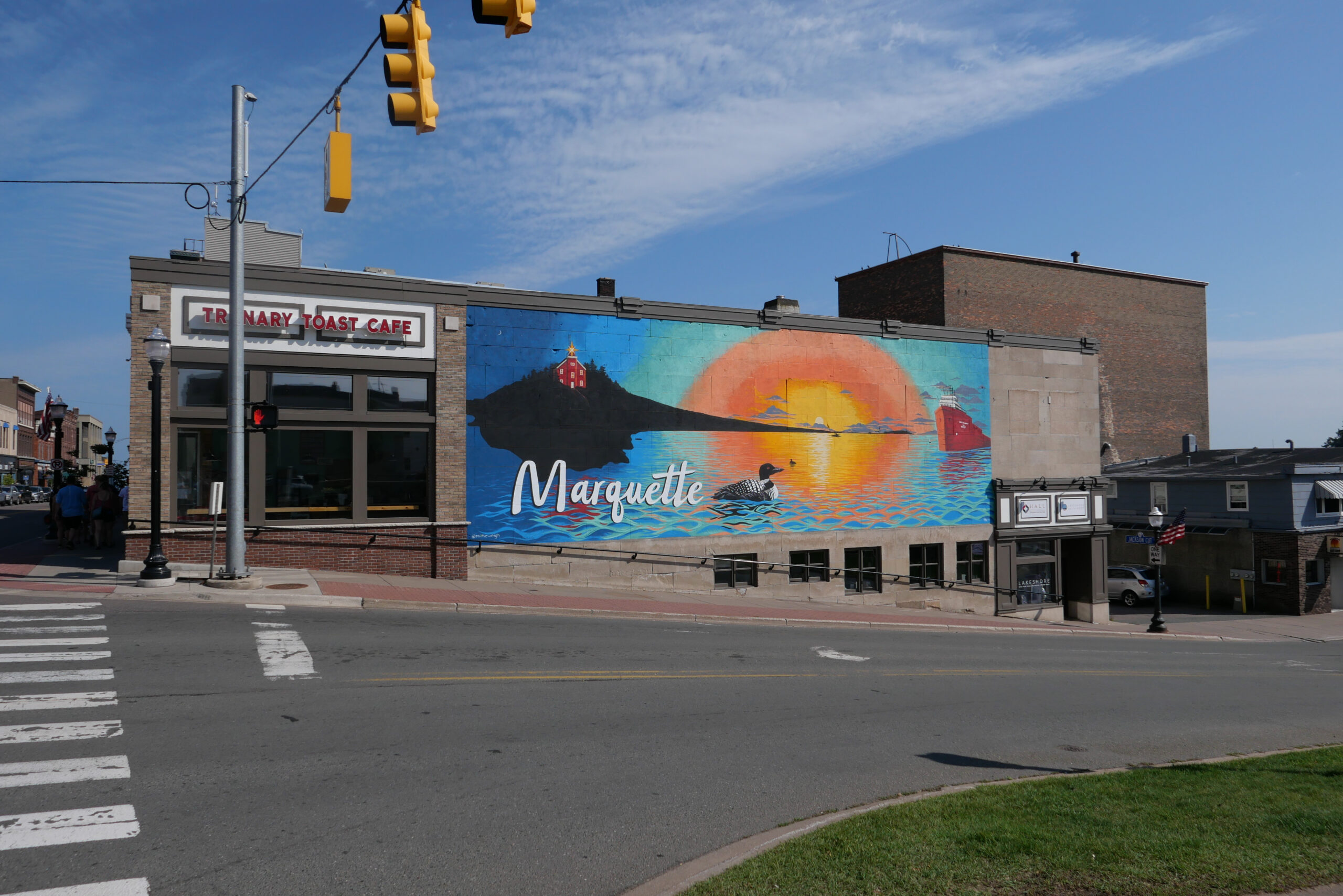A serene view of a brick building adorned with a vibrant mural on its side wall. The mural features elements like waves, the sun, and the text “Marquette.” The scene is set against a clear blue sky, with a well-marked pedestrian crossing leading towards the cafe. The building houses the “TRENARY TOAST CAFE”