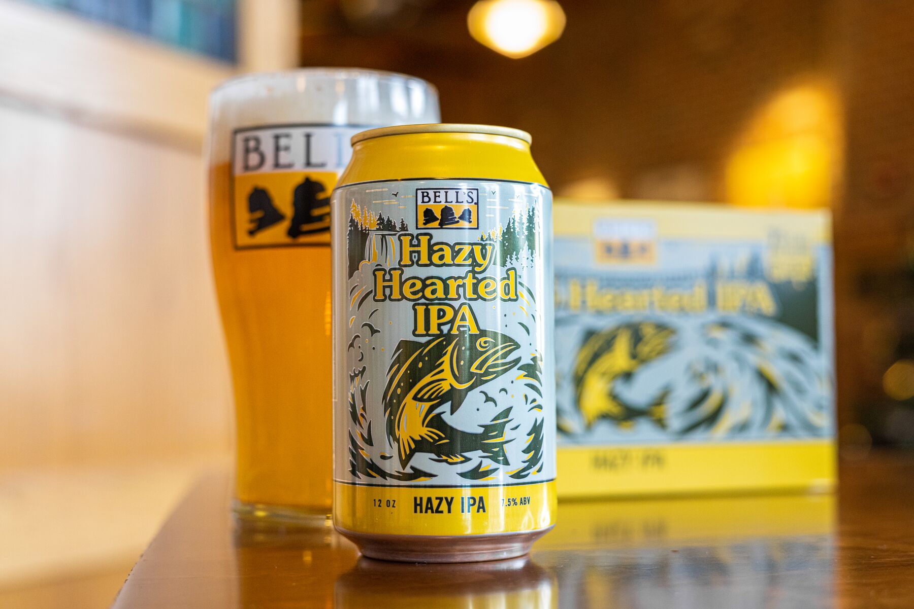 A can of Bell's Hazy Hearted IPA, in front of a pint glass with the Bell’s logo and a bright gold beer in it. The cans feature colorful artwork depicting a fish jumping out of water. The cans sit on a reflective surface, with a blurred sign or menu visible in the background. The overall lighting appears warm and inviting, suggesting a cozy bar or brewery setting.