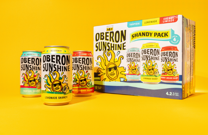 Oberon Sunshine Shandy Pack to the right with one of each can to the left (Tropical Shandy, Lemonade Shandy, Cherry Limeade Shandy) on a yellow sweep.