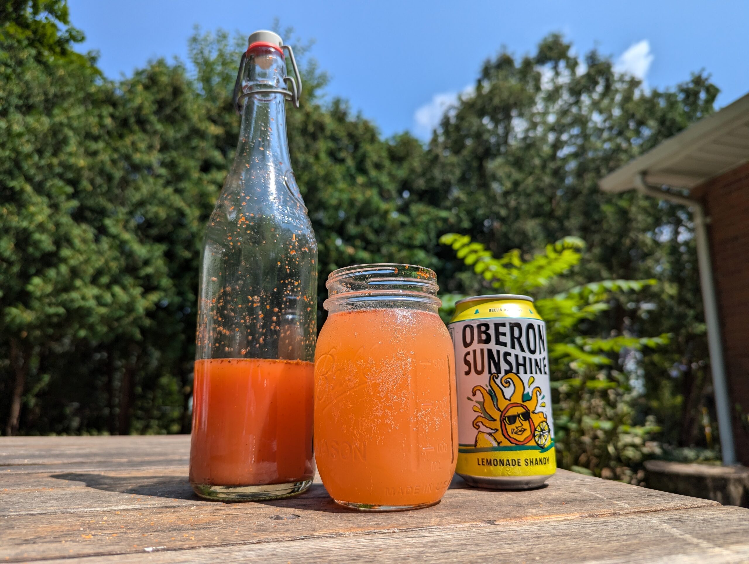 Orange cocktail in a mason jar with a bottle and an can of Oberon Sunshine Shandy next to it outside on a wooden table with trees in the background.