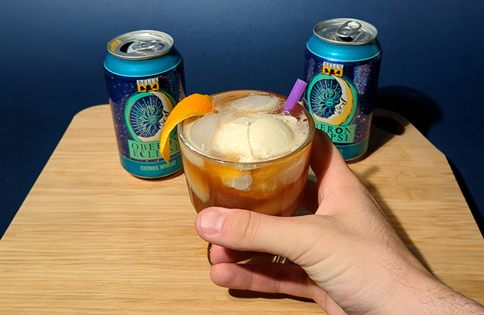 A hand is holding a glass filled with an Old Fashioned cocktail topped with Bell’s Oberon Eclipse and a scoop of vanilla ice cream. On either side of the glass are two cans of Oberon Eclipse, all set on a cutting board with a deep blue background.