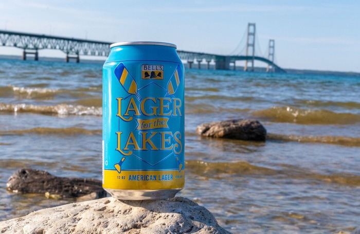 A can of Lager for the Lakes perched atop a rock with a lake and bridge in the background.